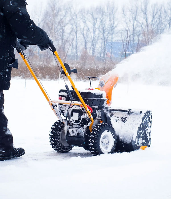 A person operates a snow blower, clearing a path through freshly fallen snow in a residential area.